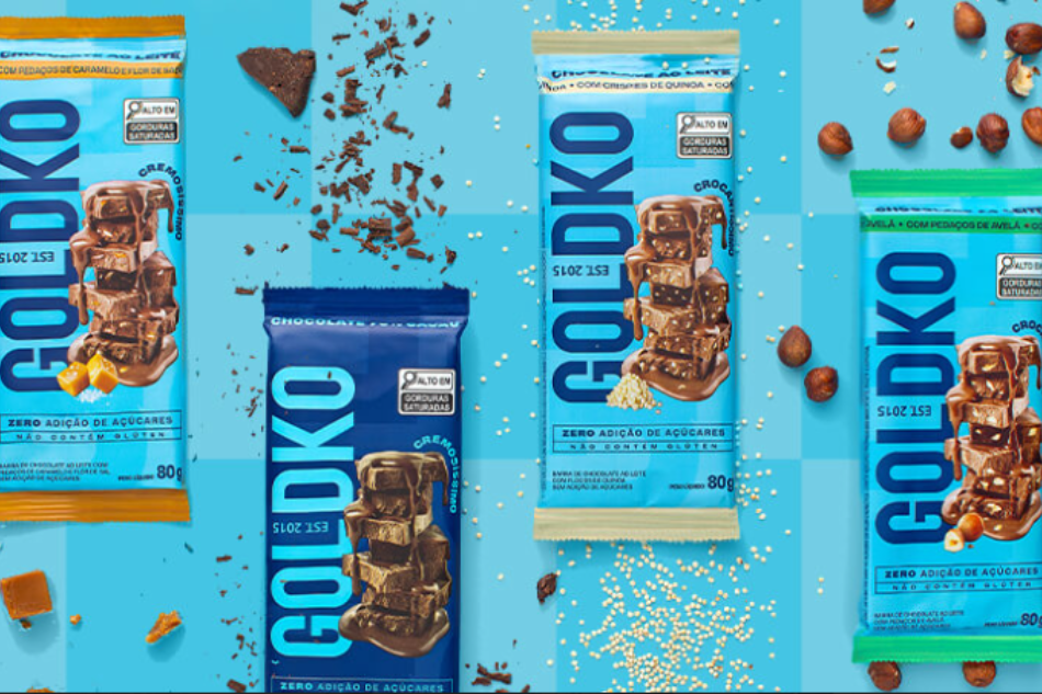 Barras de chocolate da marca GoldKo dispostas em um fundo azul, com pedações de cacau, doce de leite, avelãs e quinoas para simbolizar os recheios.